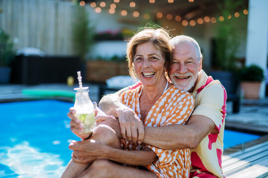 Happy senior couple enjoying drinks when relaxing and sitting by swimming pool in summer.