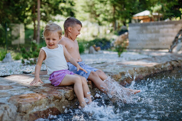 Cute girl and boy sitting together on the footpath by the lake, dangle their feet in the water, summer vacation concept.