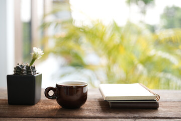 coffee brown ceramic cup and white cactus flower in a black pot on wooden table with notebook