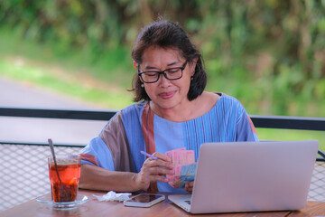 A middle-aged smiling woman is counting some money; a glass and a laptop in front her
