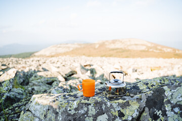 Tourist utensils stand on a stone in the mountains, an orange mug for tea, a hot drink in nature,...