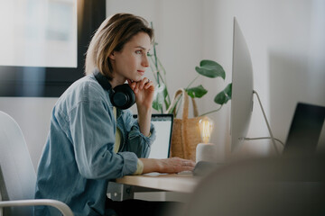 A female freelancer working from home in a well-lit room, with headphones on and focused on her...