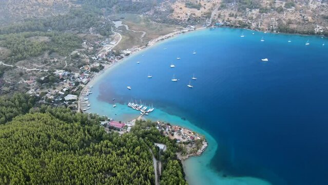 Aerial drone video showcasing the picturesque Akb&uuml;k Cove in Akyaka, Muğla, Turkey, featuring yachts and boats anchored in the pristine waters surrounded by a scenic coastline, mountains, and trees.