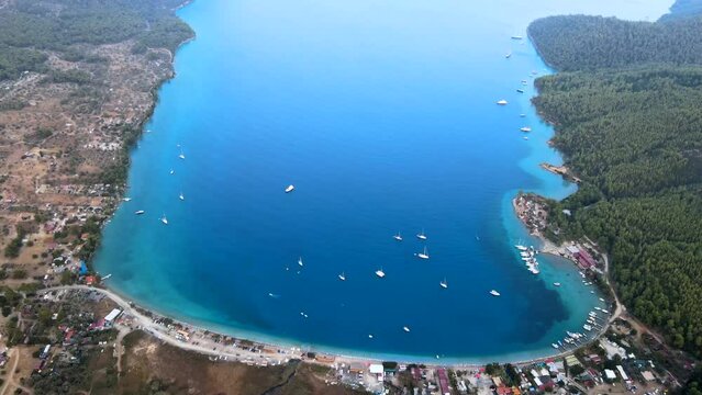 Aerial drone video showcasing the picturesque Akb&uuml;k Cove in Akyaka, Muğla, Turkey, featuring yachts and boats anchored in the pristine waters surrounded by a scenic coastline, mountains, and trees.