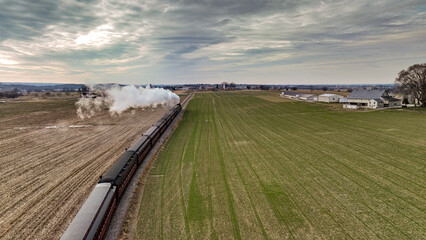 A Behind Aerial View of a Steam Passenger Train Approaching, Traveling Thru Open Farmlands, Blowing Lots of White Smoke, on a Winter Day