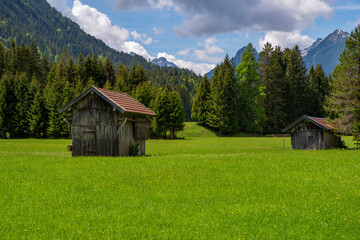 Fototapeta na wymiar View to two sheds on green meadow on a sunny day, blue sky with white clouds and mountains in background