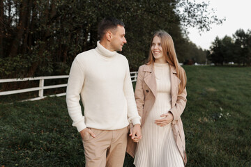 Fototapeta premium Young happy romantic pregnant couple is walking holding hands outdoors at autumn warm day. Pregnant woman in knit dress and her handsome man expecting a baby. Happy family day. Parenthood, motherhood