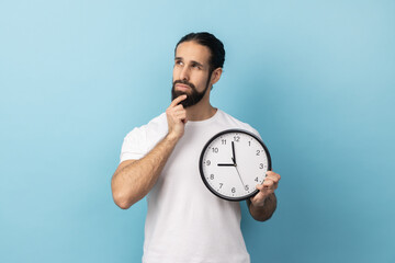 Portrait of thoughtful pensive man with beard wearing white T-shirt holding wall clock and holding his chin, deadline, punctuality. Indoor studio shot isolated on blue background. - obrazy, fototapety, plakaty