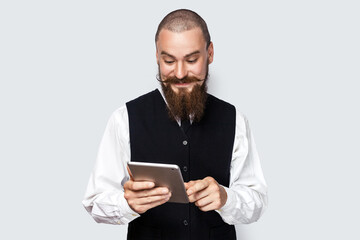 Portrait of smiling optimistic handsome man with beard and mustache standing using tablet, chatting with friends, reading message in social network. Indoor studio shot isolated on gray background.