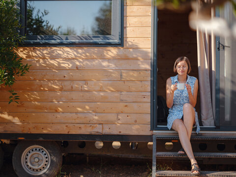 Woman is drinking coffee , morning routine near her Tiny house. First property. Small apartment in summer garden. Minimalism. Moving in. Living alone. Charming trailer house