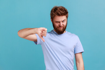Portrait of bearded man criticizing bad quality with thumbs down displeased grimace, showing dislike gesture, expressing disapproval. Indoor studio shot isolated on blue background. - obrazy, fototapety, plakaty