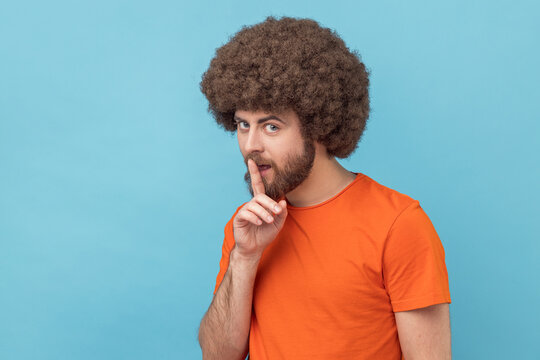 Portrait of man with Afro hairstyle wearing orange T-shirt showing hush with finger on his lips gesture, shushing, asking to keep silence. Indoor studio shot isolated on blue background.