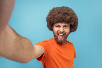 Portrait of excited positive optimistic man with Afro hairstyle wearing orange T-shirt taking selfie, expressing positive emotions, point of view photo. Indoor studio shot isolated on blue background.
