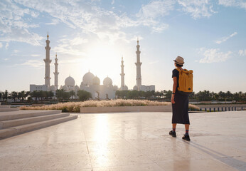 travel to the United Arab Emirates, Happy young asian female traveler with backpack and hat in Wahat Al Karama or Oasis of Dignity, permanent memorial for its martyrs, and Shaikh Zayed Grand Mosque. - obrazy, fototapety, plakaty