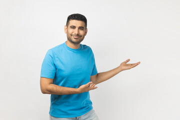 Portrait of smiling friendly happy unshaven man wearing blue T- shirt standing showing welcome gesture, inviting to come, looking at camera. Indoor studio shot isolated on gray background.