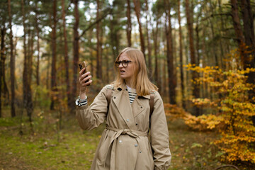 Woman in a style beige coat stands in a autumn forest on a hike and catches a phone call on a smartphone with his arm raised up. Hiker looking for a mobile network on a smartphone. High quality photo