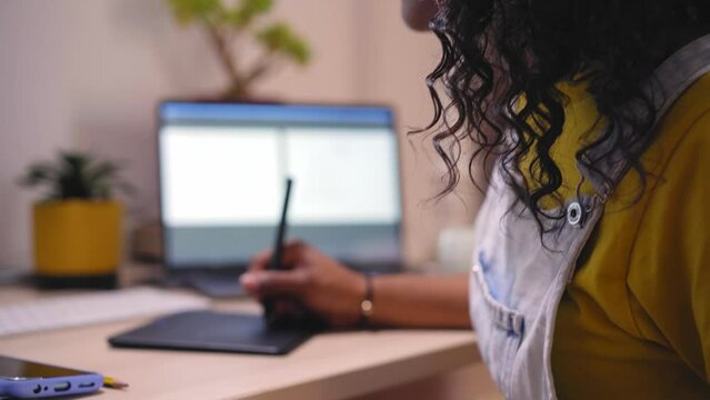 Focus shot on face and hands of an African American woman working with a drawing tablet for a computer. Freelance girl practicing from home at a desk. Young people making a living online indoors.