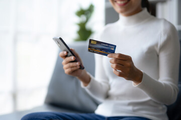 Happy young Asian woman using smartphone and credit card for online shopping on sofa at home.
