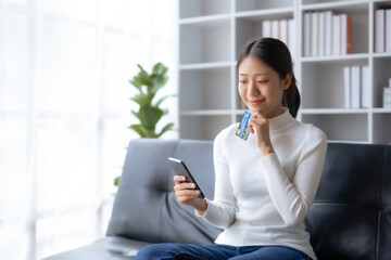 Happy young Asian woman using smartphone and credit card for online shopping on sofa at home.