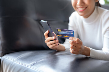 Happy young Asian woman using smartphone and credit card for online shopping on sofa at home.