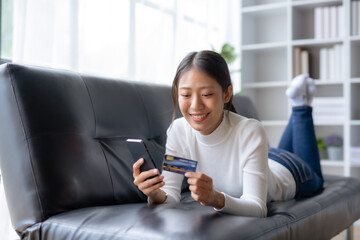 Happy young Asian woman using smartphone and credit card for online shopping on sofa at home.