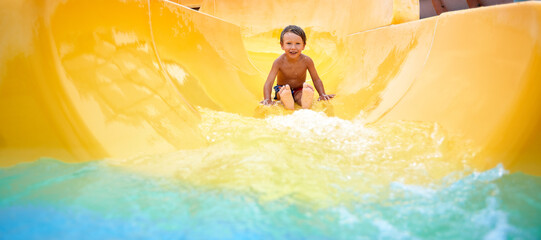 Cheerful blue-eyed kid on a water slide in the water park, a little boy merrily slides into the...