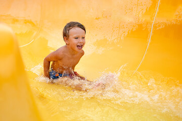 Cheerful blue-eyed kid on a water slide in the water park, a little boy merrily slides into the...