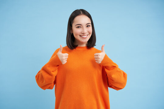 Good job. Smiling korean woman shows thumbs up in approval, like smth, gives good feedback, nods in positive reply, recommends something, blue background