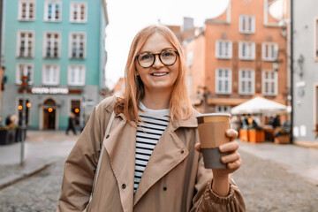 Obraz premium Young smiling woman in coat with coffee cup in evening city street in Gdansk old town. Traveling Europe in autumn. High quality photo
