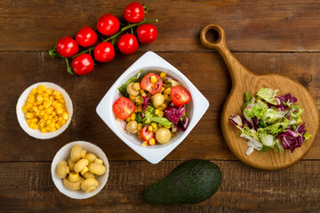 Vegetable salad with champignon corn and cherry tomato with avocado in a white plate on a wooden table next to all the ingredients.