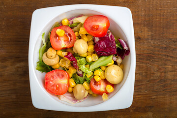 Vegetable salad with champignon corn and cherry tomato with avocado in a white plate on a wooden table.