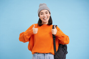 Enthusiastic asian woman in hat, holds backpack, shows thumbs up, approves smth, gives positive...