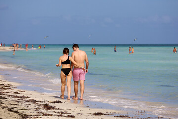 Couple walking by the sand on sea waves and swimming people background. Tropical beach with tourist resort on Caribbean islands