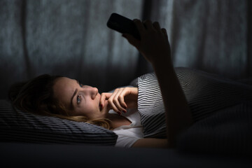 Young woman in bed holding a phone, tired and exhausted, blue light straining her eyes, messing up...