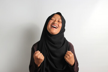 Excited young Asian girl in hijab raising fist isolated on white background.