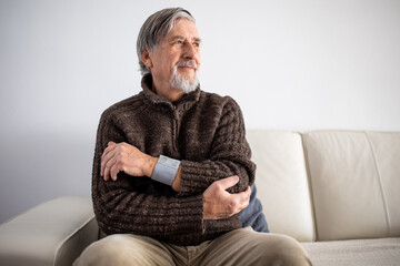 Obraz premium Senior man using medical device to measure blood pressure - elderly man suffering from high blood pressure sitting at home on sofa takes care of his health