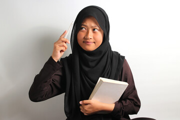 Thoughtful young Asian girl student interested in thinking while holding a book and pen isolated on white background