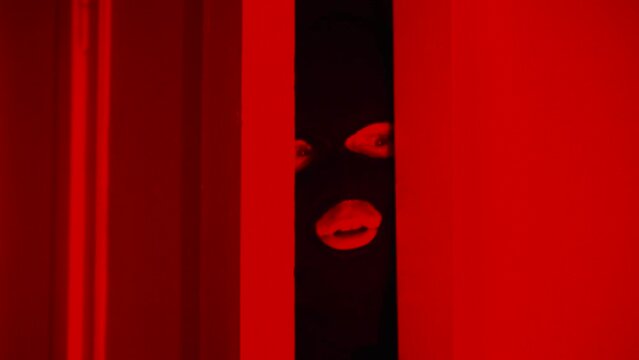 Frightened thief looks out of the door under the flashing police siren, a comical man in a thief's black mask hides from the guards