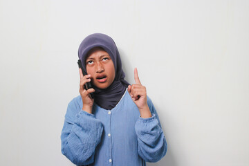 Annoyed young Asian girl in hijab dressed in casual shirt holding phone and pointing finger up