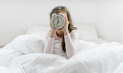8 years girl holding alarm clock 7 am hidding the face, sitting on bed under white blanket, sleep at morning. Time go to school, wake up early at home