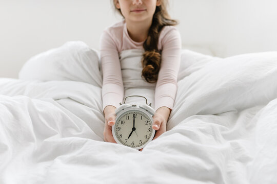 8 years girl holding alarm clock 7 am sitting on bed under white blanket, sleep at morning. Time go to school, wake up early at home