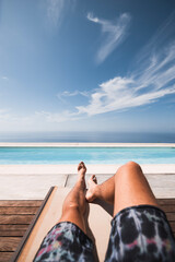 first-person photo of a man's legs sunbathing in a hammock in front of an ifinity pool with the sea in the background