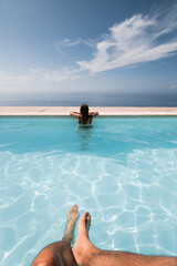 girl from behind looking at the sea inside an infinity pool with some man's legs in the foreground on a sunny day