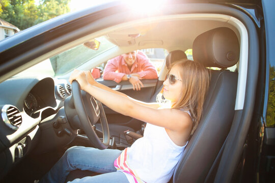 Young girl sitting in driver's seat of car, pretending to be old enough to drive as her smiling father watches on on a sunny summer evening in Portland, Oregon, USA