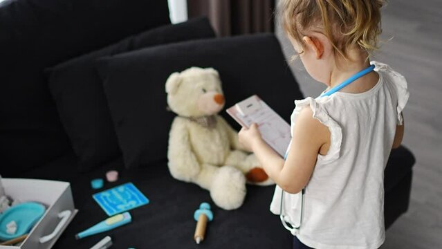 Little girl playing doctor with toys and teddy bear on the sofa in living room at home