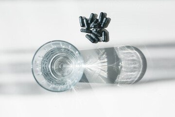 Top view of sparkling white glass with sunlight, long shadow and patterns of refraction. A glass of water with the black pills on a white table. Absorbent.
