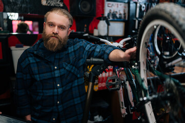 Fototapeta na wymiar Portrait of handsome bearded cycling repairman standing by bicycle in repair bike workshop with dark interior, looking at camera. Concept of professional repair and maintenance of bicycle transport.