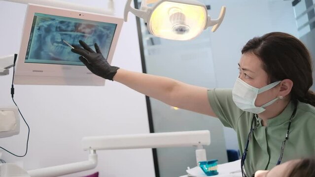 Woman dentist and patient reviewing digital X-ray on screen at modern clinic and discussing teeth treatment 