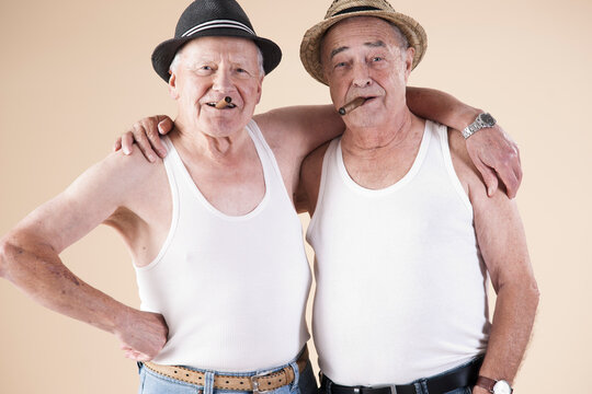 Portrait of Two Senior Man wearing Undershirts and Hats while Smoking Cigars with Arms around Shoulders, Studio Shot on Beige Background