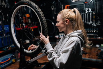 Side view of smiling blonde female cycling craftsman checking bicycle wheel spoke with bike spoke key in repair workshop with dark interior. Concept of professional maintenance of bicycle transport.